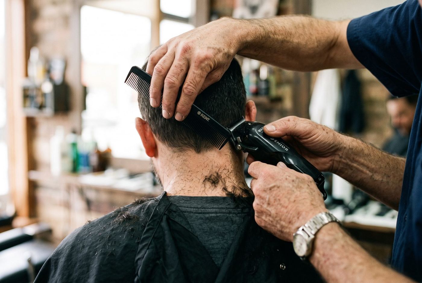 A barber steadies a man's head with one hand and runs a clipper up the back of a comb held against the hairline, cut hair scattered on the cape below.